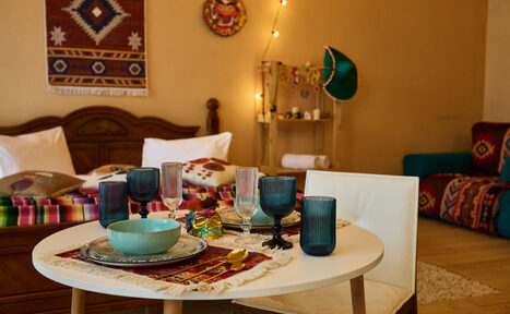 Арендовать ⁠Лофт в Мексиканском стиле для костюмированных вечеринок. Круглый белый стол с разноцветной посудой, накрытый тканью с этническим узором, рядом белый стул. На фоне кровать с подушками и покрывалом, декорированные стены, светящаяся гирлянда, полка, рядом синие и красные элементы декора, ковёр на полу.