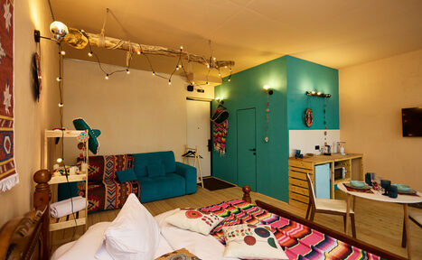 Арендовать ⁠Лофт в Мексиканском стиле для костюмированных вечеринок. Комната с кроватью, диваном и обеденным столом. Яркие текстильные элементы. Голубая акцентная стена с дверью. Настенная полка и стойка с посудой. Деревянный пол и кровать. Лампочки на проводе, подсветка над столиком. Светлая отделка, декоративные элементы.