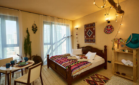 Арендовать ⁠Лофт в Мексиканском стиле для костюмированных вечеринок. Спальня с деревянным полом, двуспальная кровать с ярким покрывалом, настенные декорации, стол с двумя стульями, открытая деревянная полка с предметами, большие окна с полупрозрачными шторами, растения, подвесное освещение на ветке.