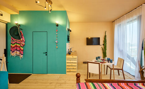 Арендовать ⁠Лофт в Мексиканском стиле для костюмированных вечеринок. Гостиная с ярко-голубыми и бежевыми стенами, деревянный пол, стол с посудой и двумя стульями, ТВ на стене, декоративные элементы, кактус, цветные покрывала и шляпы, мягкое освещение, большие окна с белыми шторами.