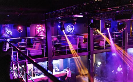 Арендовать Castle Concert Hall(Концертный зал Замок). Двухуровневое пространство с балконом, оборудованное барной стойкой. Яркое цветное освещение, пара кресел у стен с картинами. Металлические перила и конструкции. Люди за баром. Световые лучи отражаются от поверхностей.