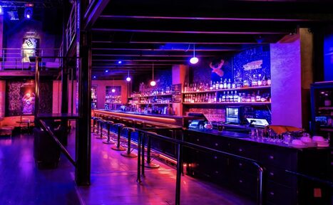 Арендовать Castle Concert Hall(Концертный зал Замок). Бар с длинной стойкой и высокими стульями, рядом стойка для микшеров. Темное освещение с неоновыми фиолетовыми и розовыми оттенками. На заднем плане полки с бутылками, стены с графикой. Потолок с балками, металлические конструкции.