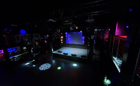 Арендовать Castle Concert Hall(Концертный зал Замок). Ночной клуб с двумя уровнями и сценой. Центральный экран, несколько световых эффектов на полу. Оборудование для света и звука. Барная стойка слева, металлические перила вдоль второго уровня. Черный интерьер с разноцветной светодиодной подсветкой.