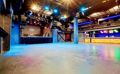 Арендовать PRAVDA(Правда). Просторный зал на 800 гостей. Большое открытое пространство с бетонным полом, сцена с музыкальным оборудованием, барная стойка с подсветкой, разноцветные прожекторы на потолке, металлические конструкции. Потолок с обширной системой освещения и проводки. На экране надпись "PRIZMA".