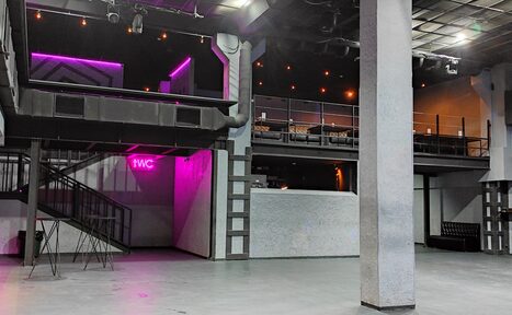 Арендовать PRAVDA(Правда). Просторный зал на 800 гостей. Многоуровневое пространство с бетонным полом, колоннами и балконом. Черная лестница ведет на второй этаж. Неоновая вывеска «WC». Тёмное освещение с точечными светильниками. Стены серые с текстурой. Мебель включает высокие стулья и диваны.