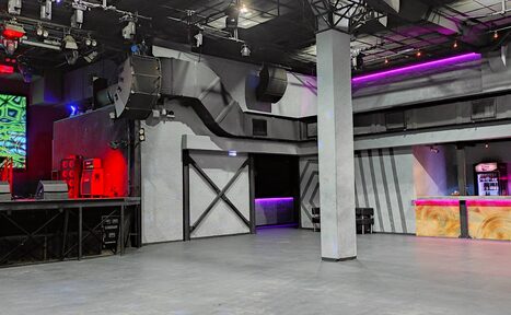 Арендовать PRAVDA(Правда). Просторный зал на 800 гостей. Клубное пространство с бетонным полом и металлическими колоннами. Справа барная стойка с декоративной подсветкой фиолетового и красного цветов. Слева сцена с аудиосистемой. Потолок с подвесными светильниками и колонками. Стены серые с черными элементами.