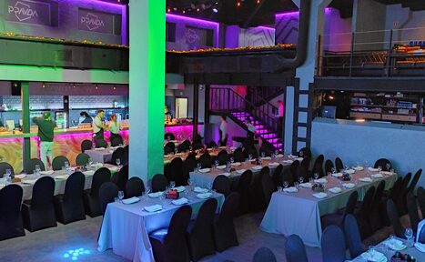 Арендовать PRAVDA(Правда). Просторный зал на 800 гостей. Просторное помещение с двумя уровнями, прямоугольные столы с белыми скатертями, стулья с тёмными чехлами, яркое разноцветное освещение, неоновые элементы, металлическая лестница, знаки с надписью "PANDA" на верхнем уровне, барная стойка с подсветкой.