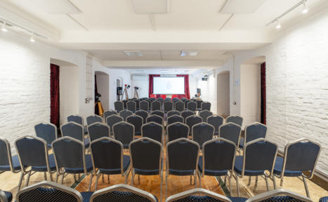 Арендовать Светлый Лофт. Пространство для мероприятий с белыми кирпичными стенами и деревянным полом. Ряды черных стульев с серыми вставками, направленных к белому экрану проектора. Потолок с точечным освещением, в задней части помещения установлены штативы для камер.