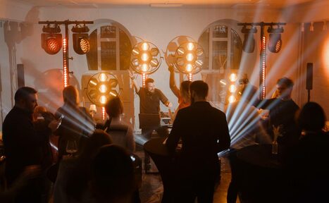 Арендовать Светлый Лофт. Танцевальная площадка с мягким освещением, круглыми столами, цветочными вазами, сценой и диджейским оборудованием. Световые прожекторы, лучи света. Интерьер в тёмных и светлых тонах, окна на заднем плане.