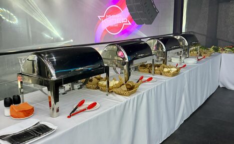 Арендовать СВОБОДА. Зал на 1200 человек. Помещение для банкета с длинным столом, покрытым белой скатертью. На столе четыре металлических контейнера для еды, подносы с хлебом, красные щипцы, посуда, посолонки и салфетки. На заднем плане светодиодные панели с текстом.