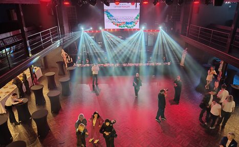 Арендовать СВОБОДА. Зал на 1200 человек. Большое внутреннее пространство с деревянным полом и балконами на втором уровне. На переднем плане высокие круглые столы. На заднем плане сцена с экраном и световыми эффектами. Люди стоят и общаются. Освещение разноцветное.