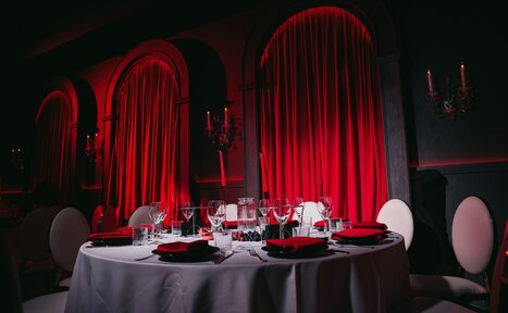 Арендовать QUQLA. Круглый стол с белой скатертью и сервировкой на шесть персон; красные салфетки, стеклянная посуда. Стулья с белой обивкой. На стенах арочные ниши с закрытыми красными шторами, красное освещение, декоративные настенные канделябры.
