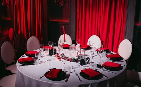 Арендовать QUQLA. Круглый обеденный стол с белой скатертью и красными салфетками, окруженный белыми стульями. На столе бокалы, тарелки, столовые приборы и свечи. Задник оформлен красными шторами. Освещение мягкое, акцент на красные и белые цвета.