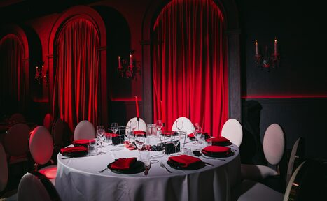 Арендовать QUQLA. Круглый стол, сервированный бокалами и столовыми приборами, окружен мягкими стульями. На столе черные тарелки с красными салфетками. Красные шторы на фоне, тёмные стены с подсвечниками. Освещение мягкое, красно-тёмная цветовая гамма.