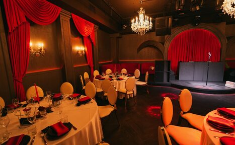 Арендовать QUQLA. Банкетный зал с круглыми столами, накрытыми белыми скатертями, сервированными бокалами и черно-красными салфетками. Стулья с кремовыми спинками. В центре сцена с микрофоном. Красные шторы, темно-коричневые стены, хрустальные люстры, мягкое освещение.