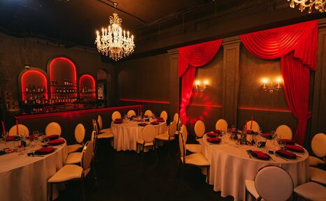 Арендовать QUQLA. Помещение для мероприятий с круглыми столами и белыми стульями. Стены украшены красными шторами и подсветкой. Пара люстр и настенные светильники обеспечивают освещение. Имеется барная стойка с полками для бутылок. Стены темно-серого цвета.