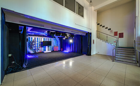 Арендовать Статус Холл. Многофункциональный зал с оборудованием. Просторное помещение с белыми стенами и плиточным полом. Сцена с черными занавесами, лампами и кабелями. Стены оформлены синим освещением. Металлическая лестница с перилами справа. Над сценой большие окна и светильники на потолке.