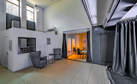 Арендовать Статус Холл. Многофункциональный зал с оборудованием. Современное многоуровневое помещение с высокими потолками. В левой части два серых кресла и столик на плиточном полу. Справа обеденная зона с круглыми столами, освещенная мягким светом. Большие окна, текстильные шторы и панели на стенах.