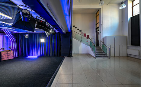 Арендовать Статус Холл. Многофункциональный зал с оборудованием. Студийное пространство с черным фоном и синим освещением, подвесное оборудование. Слева деревянный стол, справа лестница с прозрачными перилами из стекла. Пол из светлой плитки, высокие потолки, подвесные светильники.