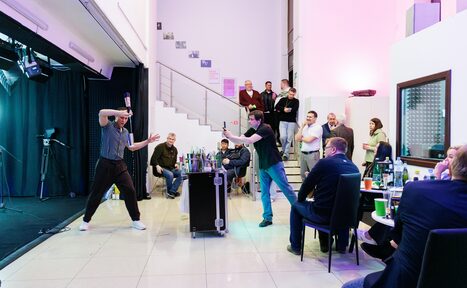 Арендовать Статус Холл. Многофункциональный зал с оборудованием. Помещение с высокими потолками, лестницей и балконом. Светлая плитка на полу. Черные кулисы и сценическое освещение. Несколько человек сидят за столами, один выступающий жонглирует бутылками. Стулья черные, столы круглые. Стены белые.