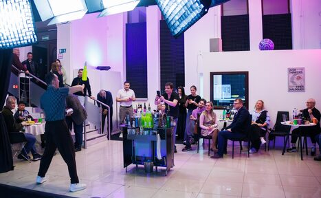 Арендовать Статус Холл. Многофункциональный зал с оборудованием. Просторное помещение с белыми стенами и плиточным полом, несколько столов и стульев с сидящими людьми, барная тележка с напитками. Яркие потолочные прожекторы, на стенах черные панели. Люди наблюдают за выступающим жонглером.