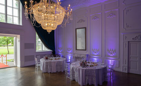 Арендовать Лофт КОСМОС с летней верандой. Просторная комната с высокими потолками; большая люстра; две круглые сервированные столы с прозрачными стульями; стены с декоративной лепниной и подсветкой фиолетового оттенка; большие окна с зелеными шторами; деревянный пол; белые двери.