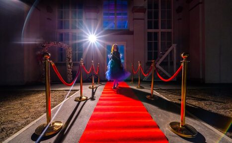 Арендовать Лофт КОСМОС с летней верандой. Красная ковровая дорожка, окруженная золотыми стойками с натянутыми красными канатами, ведет к зданию с большими окнами. Яркий прожектор освещает дорожку. Стены светлые, в углу арка украшена цветами.