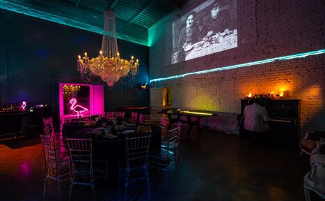 Арендовать Лофт КОСМОС с летней верандой. Большое помещение с кирпичными стенами, деревянными полами и высоким потолком. Центральная часть занята круглым столом и стульями. На стене висят неоновые вывески, большая люстра, проектор показывает изображение. Атмосферное освещение.