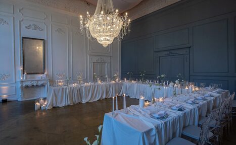 Арендовать Лофт КОСМОС с летней верандой. Зал с длинными сервированными столами с белыми скатертями, стулья из прозрачного пластика. На столах свечи и бокалы, освещение от большого подвесного люстра. Стены белого и серого цвета, декоративные панели, высокое зеркало на стене. Пол темного цвета.