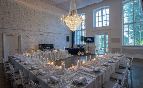 Арендовать Лофт КОСМОС с летней верандой. Большой зал с кирпичными стенами, высокой потолочной люстрой и большими окнами. Расставлены обеденные столы в П-образной форме с белыми скатертями, прозрачные стулья, свечи и сервировка. На заднем плане колонка и экран, дверь и вид на улицу.