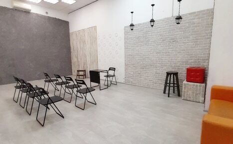 Арендовать Интер-Фото. Просторное помещение с серыми плиточными полами и белыми кирпичными стенами. Несколько рядов черных складных стульев, черный стол, высокий табурет. Ярко-оранжевое и красное сиденья. Подвесные светильники, квадратные потолочные панели.