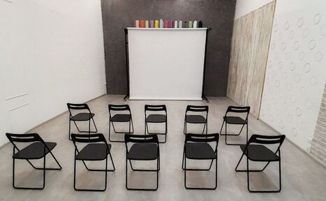 Арендовать Интер-Фото. Пространство с серыми стенами, пол из светлого материала. В центре 12 черных складных стульев, ориентированных к черно-белому экрану на штативе. Сверху экрана разноцветные цилиндры. Стена декорирована линейными и круглыми элементами.