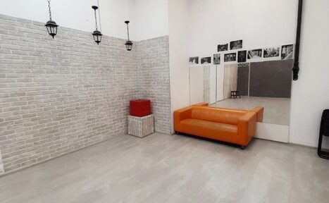 Арендовать Интер-Фото. Студийное помещение с кирпичными стенами светло-серого оттенка. Оранжевый диван, большое зеркало, чёрно-белые фотографии на стене. Лампы подвесные чёрные. Полы светло-серые. Корзина с красным покрышкой. Несколько стульев.