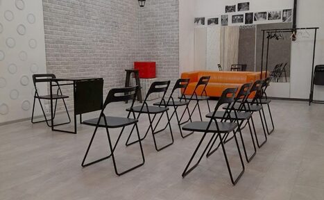 Арендовать Интер-Фото. Помещение с рядной расстановкой черных складных стульев, черный стол напротив. Оранжевый диван у стены. Белая кирпичная стена с узором из кругов и черно-белыми фотографиями. Металлическая вешалка. Светло-серый пол.