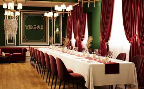 Арендовать Вегас. Атмосферный лофт с акцентным дизайном. Просторное помещение с длинным банкетным столом, покрытым белой скатертью. Красные стулья, зеленые стены, надпись "VEGAS", позолоченные светильники и красные шторы. Пол деревянный, свет из окон и люстр, украшения из сухих растений.