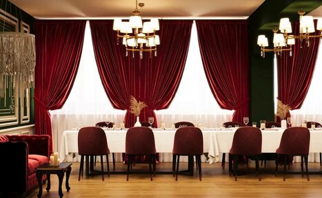 Арендовать Вегас. Атмосферный лофт с акцентным дизайном. Пространство для мероприятий с длинным столом, покрытым белой скатертью; красные бархатные занавески, стулья бордового цвета, люстры с лампами, деревянный пол, бокалы на столе, свечи на маленьком столике, зеленые стены.