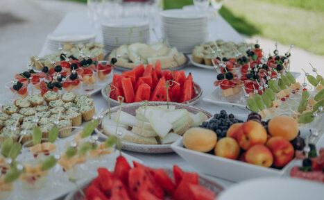 Арендовать Эко-отель "Джао Да!Ча". Шатер-Сфера. Белый стол с закусками: нарезанные фрукты, ягоды на шпажках, капкейки, десерты в стаканчиках, абрикосы, арбузы и дыни. Сложенная стопка тарелок на заднем плане. Пространство на открытом воздухе с зеленой травой.