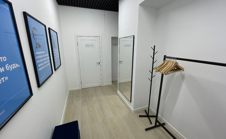 Арендовать Уютный тренинговый зал на Таганской. Узкое пространство с белыми стенами, светлым деревянным полом и черным подвесным потолком. Справа зеркало, черная стойка с вешалками и лавка. Слева три синих плаката в рамках. Две белые двери с надписями.