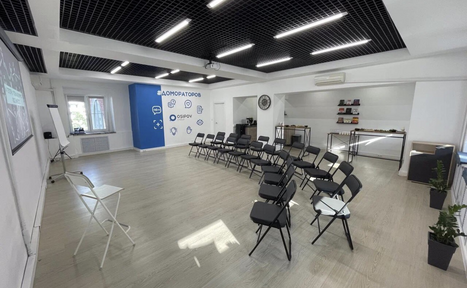 Арендовать Уютный тренинговый зал на Таганской. Просторное помещение с двумя рядами черных стульев, белыми стенами и деревянным полом. На стене виден экран и флипчарт. В правом углу столы с предметами, слева у окна кондиционер. Потолок черно-белый с подвесными светильниками.