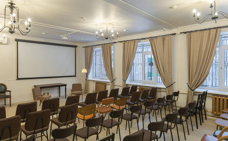 Арендовать Open Space (Открытое пространство) для мероприятий. Помещение с рядами стульев, проекторным экраном, столом и креслами. Пастельные серые и коричневые оттенки, паркетный пол. Окна с бежевыми шторами и решетками, люстры на потолке, радиаторы под окнами.