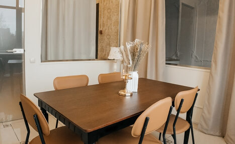 Арендовать Open Space (Открытое пространство) для мероприятий. Прямоугольный деревянный стол с шестью коричневыми стульями на металлических ножках. На столе декоративная композиция в вазе. Светлый интерьер с бежевыми шторами и светлыми стенами. Пол из плитки. Видно дверь и окно.