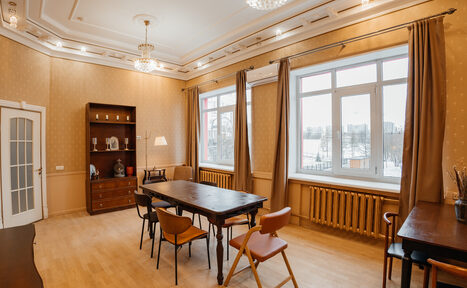 Арендовать Open Space (Открытое пространство) для мероприятий. Прямоугольная комната с высокими потолками, светлыми стенами и деревянным полом. В центре деревянный стол и стулья. Справа окно с занавесками, под ним батарея. Слева шкаф и настольная лампа. Встроенные потолочные светильники и подвесные люстры.