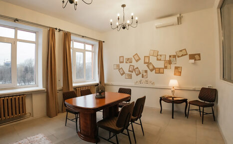 Арендовать Open Space (Открытое пространство) для мероприятий. Просторная комната с двумя окнами, столом с четырьмя стульями, двумя креслами и тумбочкой с лампой. На стене декор из пустых рамок. Светлые шторы, люстра и настенные светильники. Белые стены, бежевый пол. Радиатор под окнами. Кондиционер над картинками.