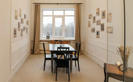 Арендовать Open Space (Открытое пространство) для мероприятий. Прямоугольная комната с высокими потолками. По центру деревянный стол и четыре стула. Светлая плитка на полу, белые стены украшены фотографиями. На переднем плане столик с вазой. Большое окно с бежевыми шторами.