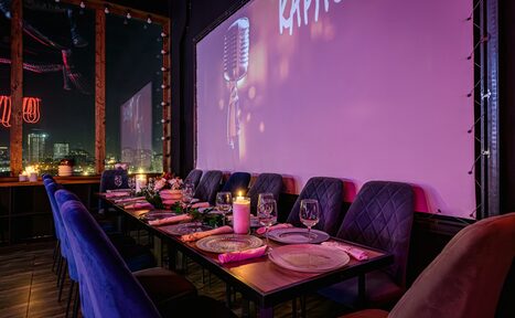 Арендовать Лофт Овертон. Просторное помещение с длинным столом, за которым стоит ряд мягких стульев. На столе сервировка с тарелками и бокалами, украшение из свечей и цветов. На стене большой экран с изображением микрофона. Темное деревянное покрытие пола, окно с видом на ночной город.