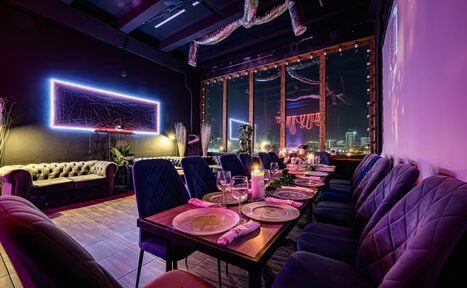 Арендовать Лофт Овертон. Лофт с деревянным полом, длинным столом, сервированным тарелками, бокалами и свечами. Вдоль стола мягкие стулья. На стене неоновая рамка. Большие окна с видом на город. Потолок с декоративными элементами. Освещение разноцветное.