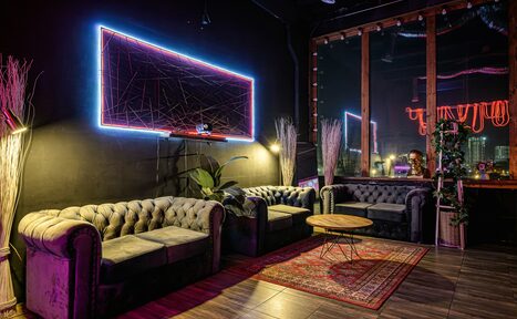 Арендовать Лофт Овертон. Лофт-пространство с темными стенами и неоновым освещением. Расположены три кожаных дивана серого цвета, стеклянный столик, деревянный пол, ковер. Витринное окно с видом на город, растительность, декоративные элементы. Светодиодная панель на стене.