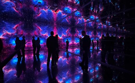 Арендовать Futurione(Футурио́не). Зеркальная комната с космическими проекциями на стенах и потолке, c яркими фиолетовыми и голубыми цветами. На полу зеркальная поверхность, отражающая изображение. Несколько людей стоят и смотрят вокруг. Освещение плавное и многоцветное.