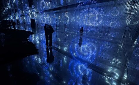 Арендовать Futurione(Футурио́не). Темное помещение с отражающим полом, стенами с нависающими нитями, подсвеченными синим светом. На полу и стенах видны узоры в виде спиралей. Несколько человек в пространстве, отражаются на полу.