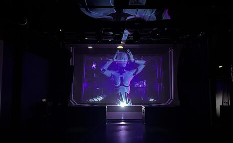 Арендовать Futurione(Футурио́не). Тёмное пространство с большой видеопроекцией, изображающей робота на экране с фиолетовым освещением. Чёрные стены и потолок. На потолке видны световые приборы и конструкции. Пол также тёмный.